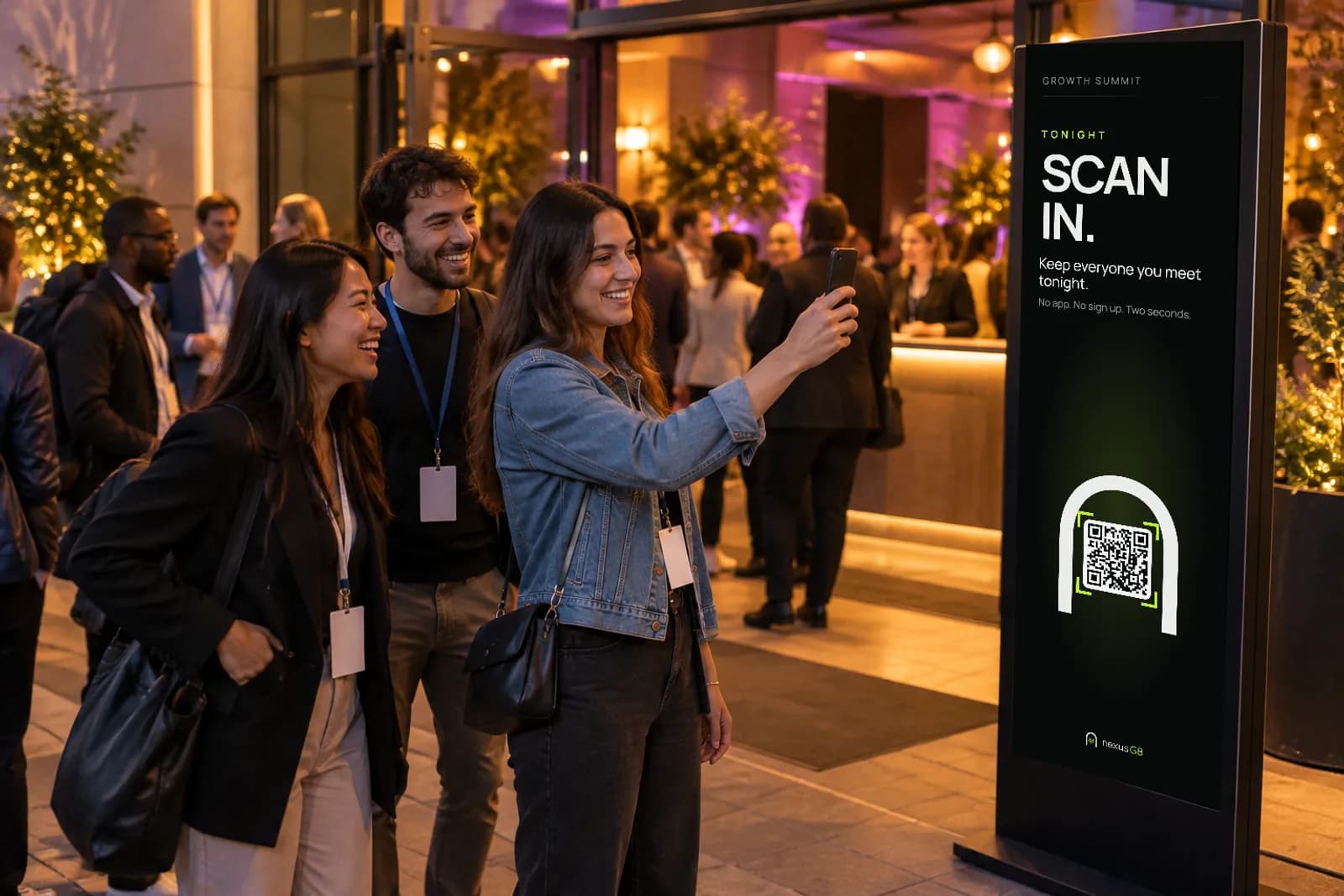At the entrance of a conference evening a woman holds her phone up to a tall screen reading Scan in, Keep everyone you meet tonight, with a NexusG8 code inside a white arch, as her friends laugh beside her